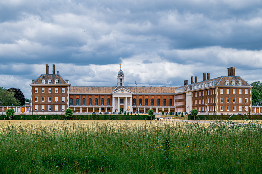 The image depicts a large, historic building with classical architecture, featuring red brick walls, white accents, and a prominent central entrance with a dome and columns. The building is set in an open field with tall grass in the foreground and a cloudy sky overhead.