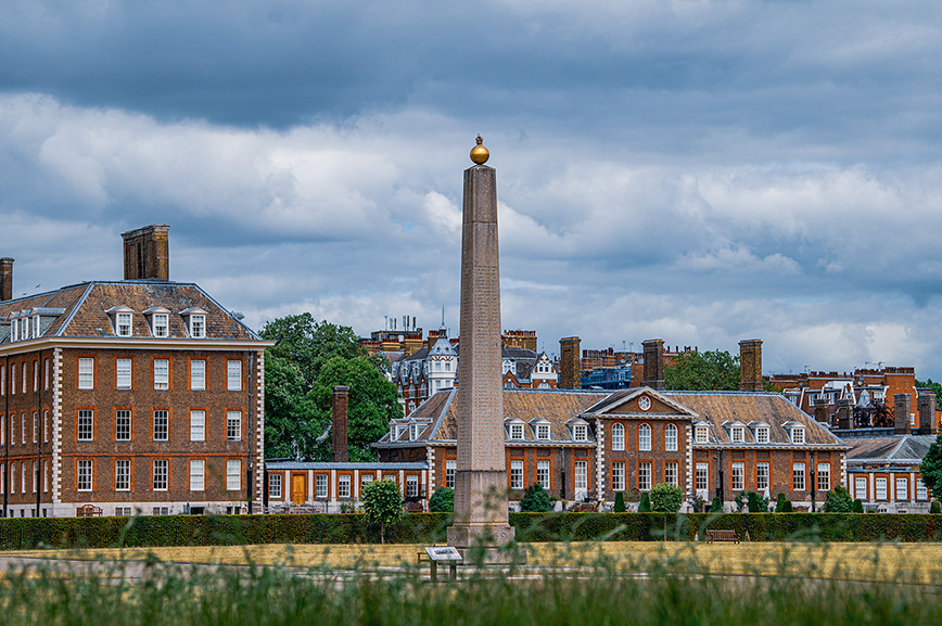 The image depicts a historic building with a tall obelisk in the foreground. The building is made of red brick with white detailing and features multiple chimneys and dormer windows. The sky is overcast, and the scene is set in a well-maintained, green open space.