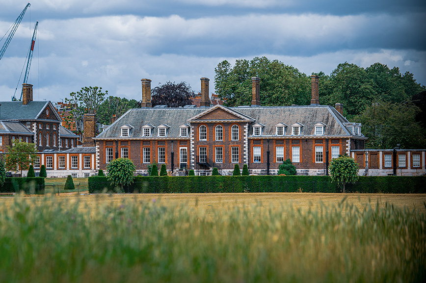 The image depicts a large, historic building with classical architecture, featuring red brick walls, white accents, and a prominent central entrance with a dome and columns. The building is set in an open field with tall grass in the foreground and a cloudy sky overhead.