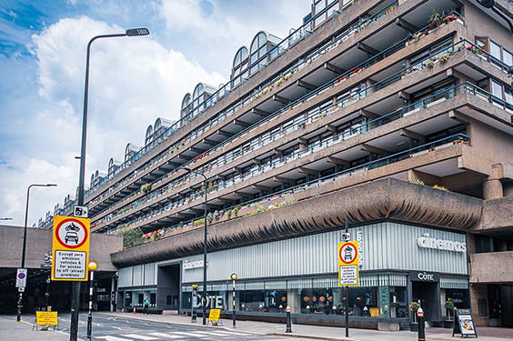 The image depicts a multi-story residential building with a distinctive architectural design, featuring multiple levels of balconies. The building is situated in an urban area with various street signs and commercial establishments on the ground floor. The sky is partly cloudy, and the overall scene suggests a typical day in a city environment.