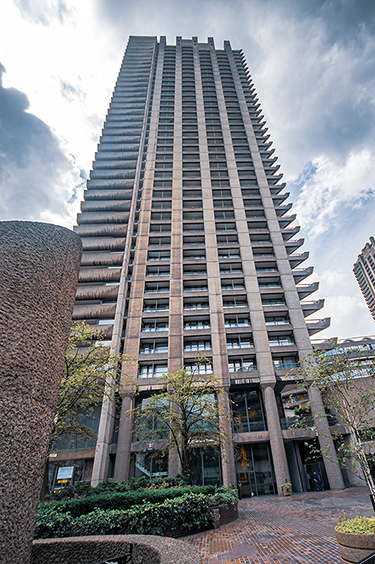 The image depicts a tall, modern high-rise building with a distinctive architectural design. The building features numerous balconies and a facade composed of a mix of concrete and glass elements. The structure is surrounded by a well-maintained garden area with greenery and a paved walkway in the foreground. The sky above is partly cloudy, and the overall scene suggests an urban setting.