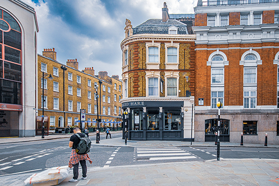 The image depicts a street scene in an urban area with a mix of architectural styles. There is a prominent corner building with ornate details and a sign that reads 'TO HARE.' The street is relatively quiet with a few pedestrians, including a person carrying a child and wearing a backpack, waiting to cross the street at a marked crosswalk. The buildings are primarily made of brick, and the sky is partly cloudy.