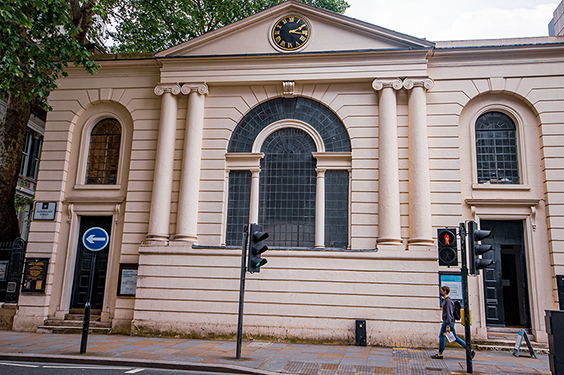 The image depicts a neoclassical building with architectural features such as columns, an arched window, and a clock on the pediment. The building is situated at a street corner with traffic lights and a pedestrian crossing. A person is seen walking past the building.