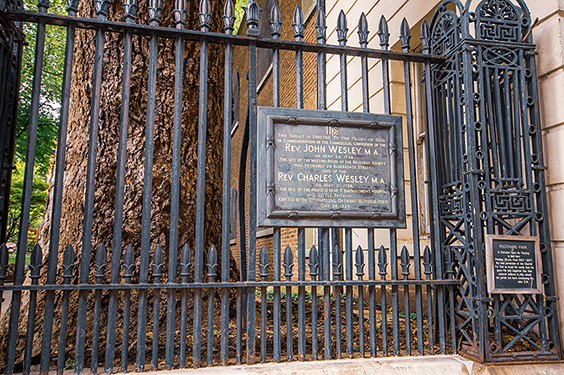 The image shows a metal gate with two plaques mounted on it. The plaques commemorate Rev. John Wesley, M.A., and Rev. Charles Wesley, M.A., noting their significant contributions and roles in religious and educational fields. The gate is part of a historical site, likely a church or educational institution, and is surrounded by trees.