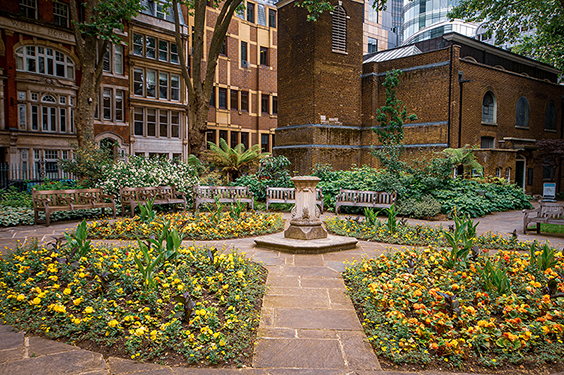 The image depicts a serene garden courtyard surrounded by multi-story buildings. The garden features a stone fountain at its center, surrounded by well-maintained flower beds and greenery. There are several benches placed around the garden, providing seating areas for visitors. The architecture of the surrounding buildings includes brick facades and large windows, contributing to a peaceful urban setting.