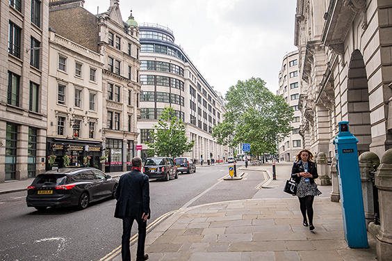 The image depicts a street scene in an urban area with several buildings, cars, and pedestrians. The architecture is a mix of older, historic buildings and more modern structures. The street is relatively quiet with a few vehicles and people visible. There is a notable blue public telephone box on the right side of the image, a common sight in the UK. The overall atmosphere suggests a typical day in a bustling city.