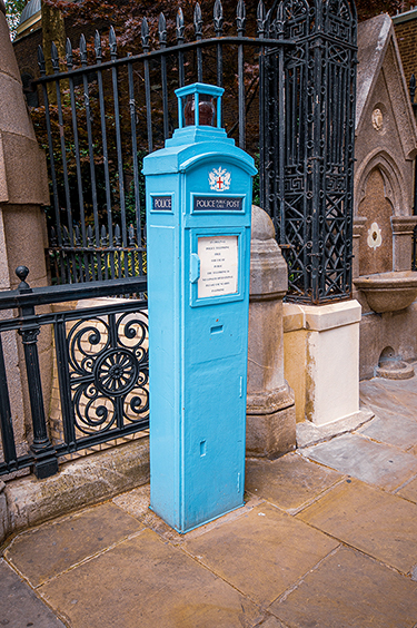 The image shows a blue police public call box situated outdoors, likely in a historical or urban setting. The box is positioned next to a black wrought iron fence and a stone wall with architectural details. There is a notice attached to the front of the box, indicating it is still in use for emergency purposes.