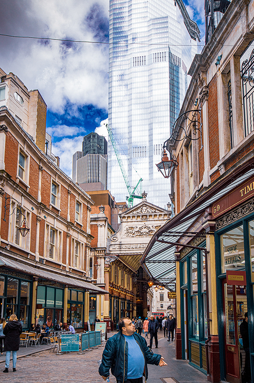 The image depicts a bustling urban street scene with a mix of modern and historic architecture. A tall, reflective skyscraper dominates the background, while older, brick buildings line the street. The street is lively with people walking and sitting at outdoor cafes. The sky is partly cloudy, adding a dynamic element to the scene. The overall atmosphere is vibrant and busy, capturing the essence of city life.