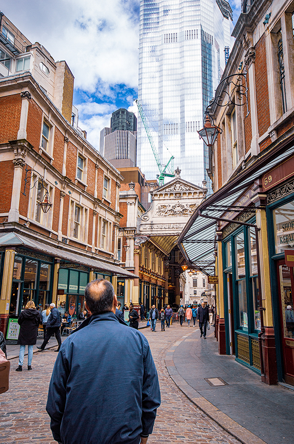 The image depicts a bustling urban street scene with a mix of historic and modern architecture. A man in a blue jacket is seen walking away from the camera, surrounded by various shops and buildings. The street is lined with brick buildings featuring ornate details and a covered walkway. In the background, tall modern skyscrapers tower over the scene, creating a contrast between old and new architectural styles. The sky is partly cloudy, adding to the vibrant atmosphere of the city.