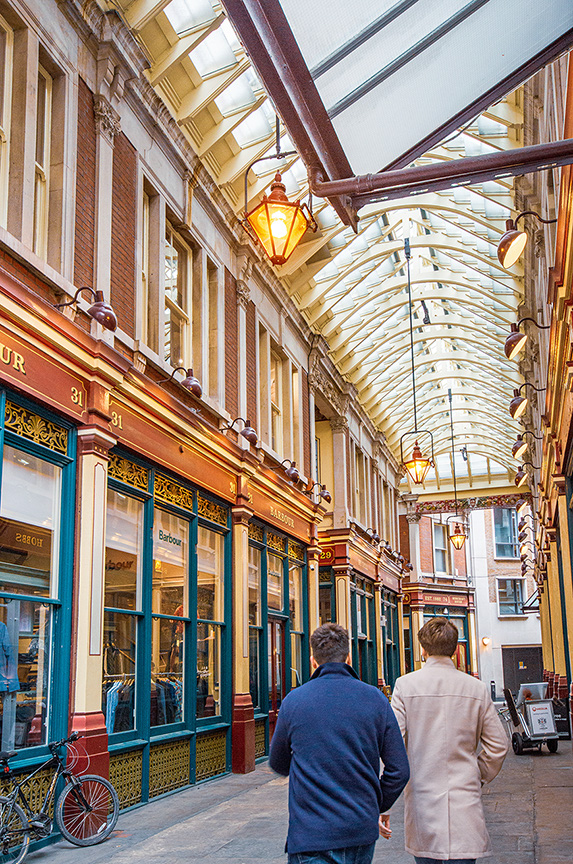 The image depicts an indoor shopping arcade with a glass-roofed ceiling, featuring two men walking away from the camera. The arcade has a vintage architectural style with ornate details, large windows displaying various shops, and hanging lantern-style lights. The overall atmosphere is bright and inviting, suggesting a pleasant shopping environment.