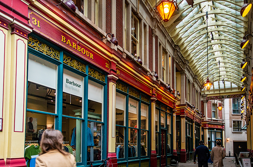 The image depicts a street view of a shopping arcade with a glass-covered walkway. The prominent store in the image is 'Barbour' with its distinctive red and gold signage. The storefronts are adorned with large windows displaying various items, and the arcade is illuminated by hanging lanterns. People are seen walking and shopping in the arcade.
