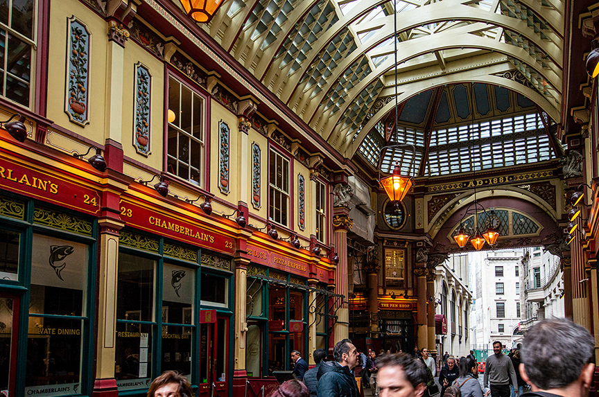 The image depicts a bustling indoor market or arcade with a high, arched glass ceiling allowing natural light to illuminate the space. The architecture features ornate details, including decorative columns and intricate moldings. Various shops line the sides of the arcade, with visible signage such as 'Chamberlain's' and 'The Pen Shop.' The area is crowded with people, indicating a lively atmosphere.