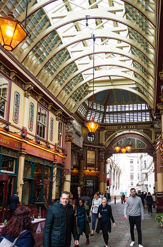 The image depicts a bustling indoor market or arcade with a high, arched, glass-paneled ceiling allowing natural light to illuminate the space. The architecture features ornate details and hanging lanterns. Various shops and eateries line the sides, with people walking through the corridor, engaging in activities such as shopping and dining.