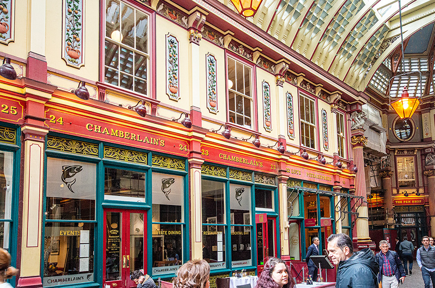 The image depicts a vibrant and ornate indoor market or shopping arcade. The architecture features colorful, detailed facades with large windows and decorative elements. The establishment named 'Chamberlains' is prominently displayed, offering events and estate dining. The arcade is bustling with people, and the ceiling is adorned with a glass roof, allowing natural light to illuminate the space.