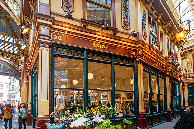 The image depicts a storefront located at 26 Royal Arcade, with the store name 'Reiss' prominently displayed above the entrance. The building features ornate architectural details, including decorative columns, intricate moldings, and large windows that allow natural light to illuminate the interior. The storefront is adorned with a variety of colorful flowers, adding a vibrant touch to the scene. People can be seen walking by and entering the store, indicating a bustling environment. The overall atmosphere suggests a lively shopping area with historical charm.