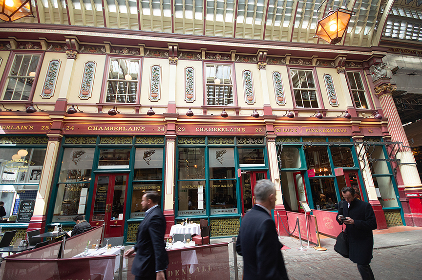 The image depicts a street scene with a row of shops and restaurants. The buildings have ornate facades with large windows and decorative elements. The establishments include Chamberlain's, Luc's, and Pizza Express. There are people walking on the street, and some are standing near a table with items on it. The overall atmosphere suggests a busy, urban setting with a mix of commercial activity.