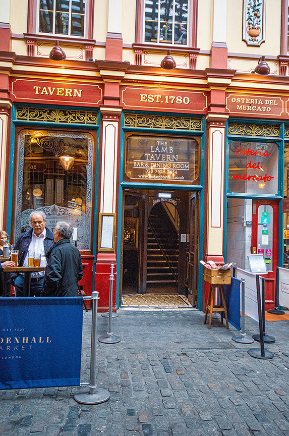 The image depicts the exterior of The Lamb Tavern, a historic establishment founded in 1780. The building features classic architecture with ornate details and signage indicating the tavern's name and contact information. Adjacent to it is Osteria del Mercato, another dining establishment. Two individuals are seen standing outside, engaged in conversation near a high table with drinks. The scene is set on a cobblestone street, adding to the historic ambiance.
