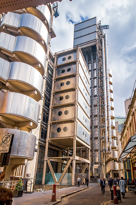 The image depicts a modern, uniquely designed building with a distinctive architectural style. The structure features a combination of cylindrical and rectangular forms, with a series of circular windows and a tall, slender tower extending upwards. The building is situated in an urban environment, with people walking on the sidewalk in front of it. The sky is partly cloudy, and the overall atmosphere appears to be bustling and lively.