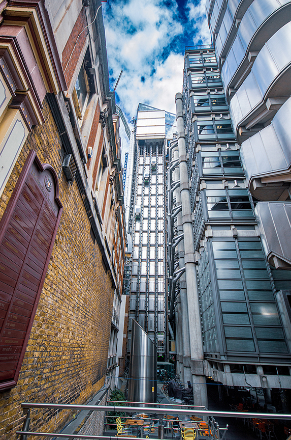 The image depicts a narrow urban alleyway flanked by tall buildings. On the left, there is an older, ornate brick building with intricate architectural details. On the right, there is a modern, glass-and-steel high-rise building. The alleyway leads to a larger, more prominent building in the background, characterized by a distinctive, industrial design with exposed pipes and a grid-like facade. The sky above is partly cloudy, adding a dramatic effect to the scene.