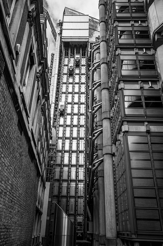 The image depicts a black and white photograph of a tall, modern building with a unique architectural design. The building features a grid-like facade with numerous windows and external structural elements. The photograph is taken from a narrow alleyway, emphasizing the height and grandeur of the structure. The surrounding buildings are more traditional in design, contrasting with the contemporary style of the central building.