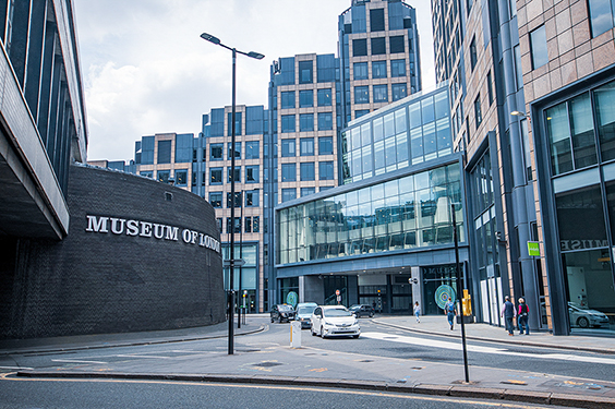 The image depicts an urban scene with the Museum of London building prominently displayed on the left. The museum has a modern architectural design with a curved facade and large glass windows. Adjacent to the museum are tall, contemporary office buildings with a mix of glass and brick exteriors. The street in front of the buildings is relatively quiet, with a few pedestrians and vehicles visible. The overall atmosphere is that of a bustling city center with a blend of cultural and commercial elements.