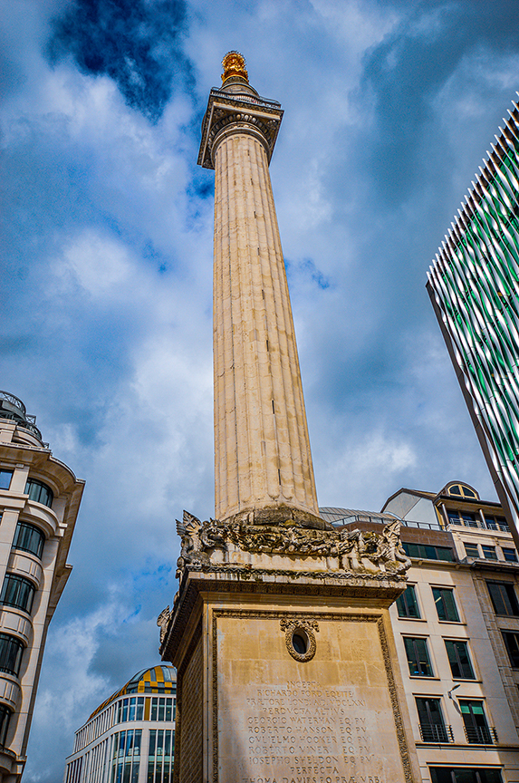 The image depicts a tall, ornate column monument set against a partly cloudy sky. The column is surrounded by modern buildings, suggesting it is located in an urban area. The base of the column features intricate carvings and inscriptions, indicating it may be a historical or commemorative structure.