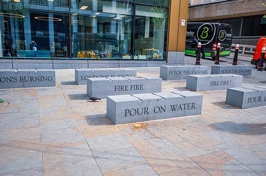 The image shows a series of concrete blocks arranged on a paved area, each block containing a segment of a message related to a fire emergency. The blocks collectively spell out: 'LONDON'S BURNING, LONDON'S BURNING, FETCH THE ENGINES, FETCH THE ENGINES, FIRE FIRE, FIRE FIRE, POUR ON WATER.' This is a reference to the old nursery rhyme and cry of fire. The scene is set in an urban environment with modern buildings and vehicles in the background.
