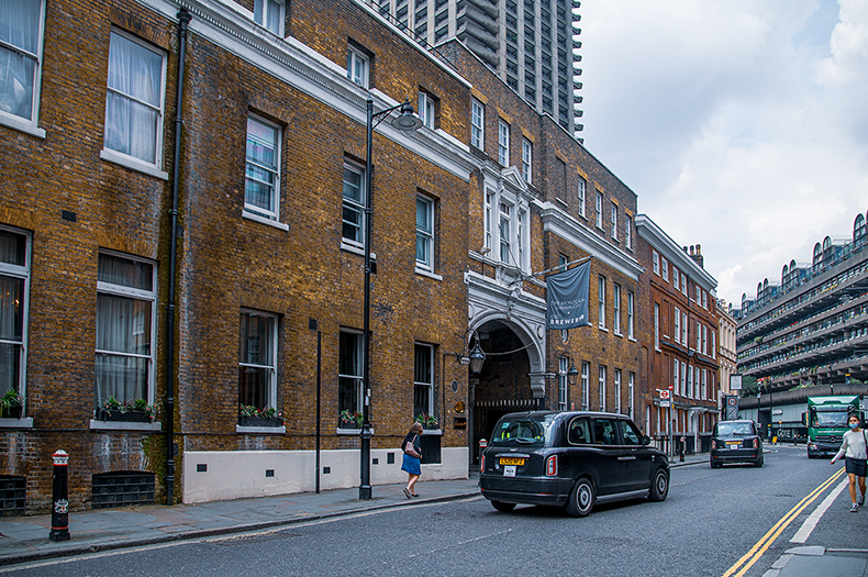 The image depicts a street scene in an urban area with brick buildings, a black taxi, and a pedestrian crossing the street. The architecture features traditional brickwork with white window frames, and there is a notable archway on one of the buildings. The scene appears to be in a city, likely in the UK, given the style of the taxi and the architecture.