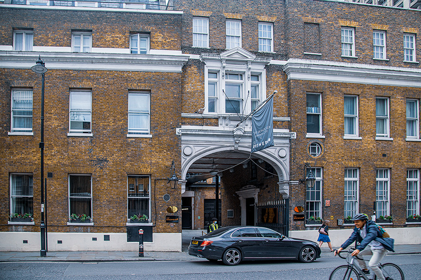 The image depicts the exterior of a brick building with a central entrance archway. The building has multiple windows with white frames and some have flower boxes. There is a flag above the entrance displaying the name 'The Resident Victoria'. A black car is parked in front of the entrance, and a cyclist is riding past on the street. The scene appears to be in an urban setting with typical city activity.