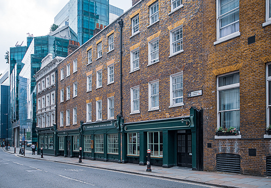 The image depicts a street scene with a row of traditional brick buildings. The buildings have large windows and some have green awnings. The street is relatively quiet with a few pedestrians and no visible vehicles. Modern glass buildings are visible in the background, indicating a blend of old and new architecture. The overall atmosphere is calm and typical of an urban setting.