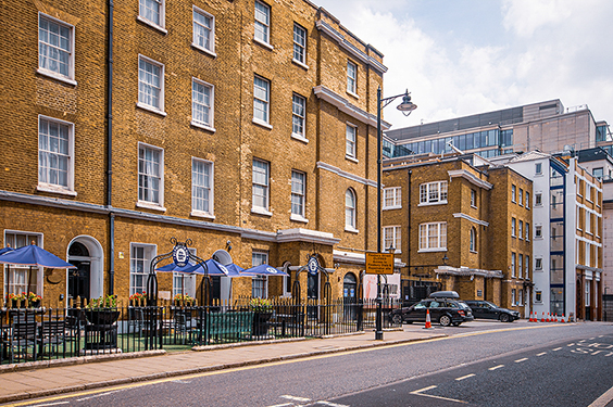 The image depicts a street scene in an urban area with a row of three-story brick buildings. The buildings have a classic architectural style with large windows and decorative cornices. Each building has a black wrought iron fence in front, and there are blue umbrellas and outdoor seating areas at the ground level, suggesting the presence of cafes or restaurants. The street is relatively quiet with a few parked cars and a single car driving by. The sky is overcast, indicating it might be a cloudy day.