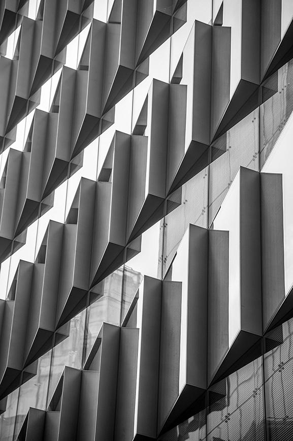 The image depicts a modern building facade with a unique geometric design, featuring a series of angular, overlapping panels that create a dynamic and intricate pattern. The structure appears to be made of metal or a similar material, with a mix of solid and semi-transparent elements. The design gives the building a contemporary and artistic appearance, with shadows and light playing off the various angles and surfaces.