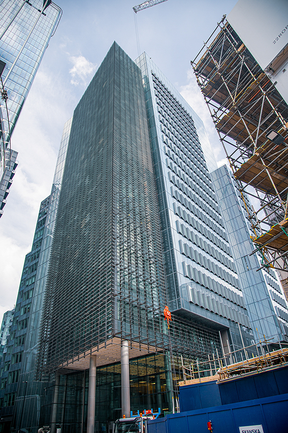 The image depicts a tall, modern skyscraper under construction. The building features a glass facade with a grid-like pattern and is surrounded by scaffolding and construction equipment. The sky is partly cloudy, and there are other high-rise buildings in the vicinity.