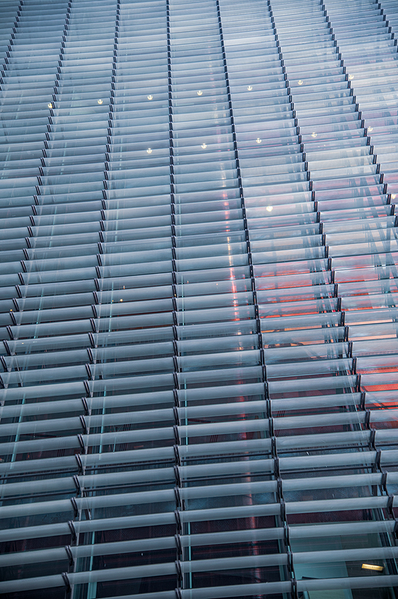 The image shows a modern building facade with a unique design featuring horizontal glass louvers. The louvers are arranged in a staggered pattern, creating an interesting visual effect. The glass reflects the sky and surrounding environment, adding a dynamic element to the building's appearance. The overall design is sleek and contemporary, emphasizing clean lines and a minimalist aesthetic.