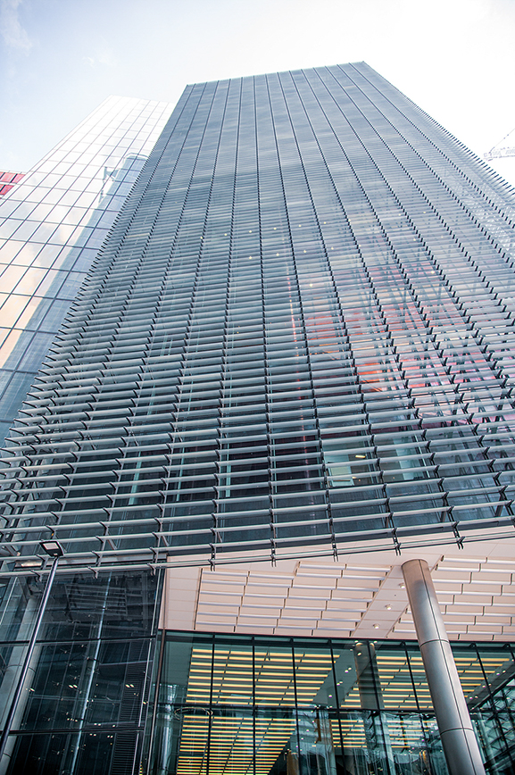 The image depicts a modern skyscraper with a distinctive facade featuring horizontal and vertical lines. The building has a sleek, glass exterior with a grid-like pattern of metal supports. The perspective of the photo is from a low angle, looking upwards, emphasizing the height and grandeur of the structure. The sky is visible in the background, suggesting it is a clear day. The architectural design is contemporary and impressive, showcasing advanced engineering and aesthetic appeal.