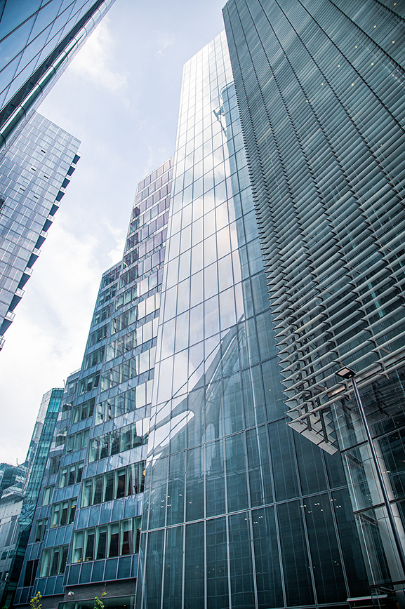 The image depicts a cluster of modern skyscrapers with reflective glass facades. The buildings are closely packed, showcasing a variety of architectural designs and reflective surfaces that mirror the sky and surrounding structures. The perspective is from a low angle, emphasizing the height and grandeur of the buildings. The sky is visible in the background, with some clouds scattered across it. The overall scene is a typical representation of a contemporary urban landscape.