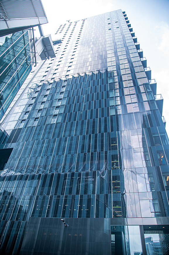 The image shows a tall, modern skyscraper with a reflective glass facade. The building has a sleek design with numerous windows and a grid-like pattern. The photograph is taken from a low angle, emphasizing the height and grandeur of the structure. The sky is visible in the background, and the reflection of nearby buildings can be seen on the glass surface.