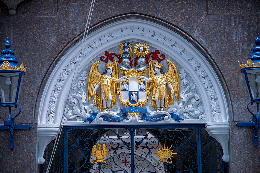 The image depicts an ornate coat of arms above a gate. The coat of arms features two golden winged figures flanking a central shield with various symbols, including a sun and a crown. The gate itself is decorated with intricate ironwork and topped with blue and gold accents. The surrounding architecture includes detailed stone carvings and decorative elements, suggesting a historical or significant building.