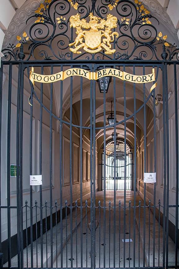 The image shows an ornate gate with a crest and the inscription 'TO GOD ONLY BE ALL GLORY'. The gate is part of an arched entrance leading to a corridor with additional gates at the far end. The architecture and design suggest a formal or historical setting.