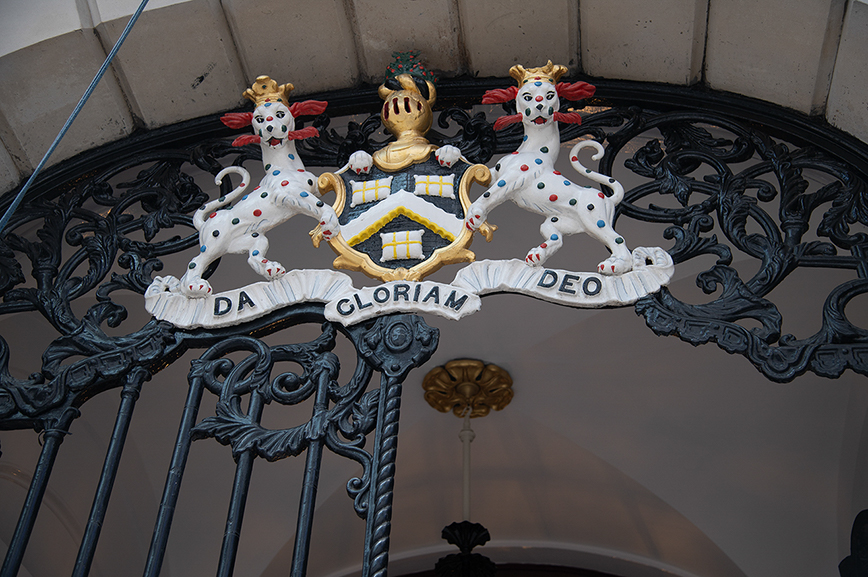 The image depicts a heraldic crest featuring two white dogs with colorful spots, adorned with red collars and gold crowns. The crest includes a shield with a gold background and blue crosses, flanked by the Latin motto 'DA GLORIAM DEO', which translates to 'Give Glory to God'. The crest is mounted on an ornate, black wrought-iron railing.