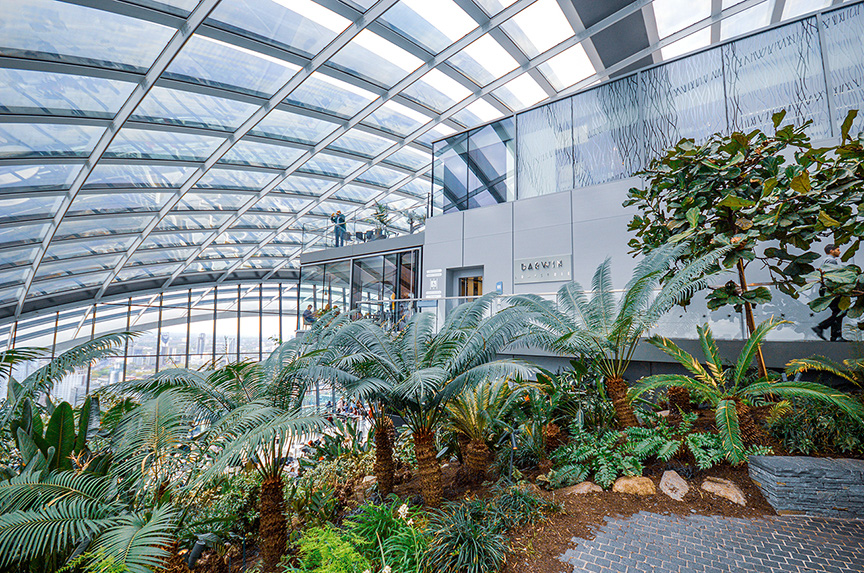 The image depicts a large indoor botanical garden with a high, glass ceiling allowing natural light to illuminate the space. The garden is filled with a variety of lush, green plants and trees, creating a tropical atmosphere. There are pathways for visitors to walk through and enjoy the greenery. The structure appears to be part of a modern building with a glass facade, and there are people visible in the background, indicating it is a public space.