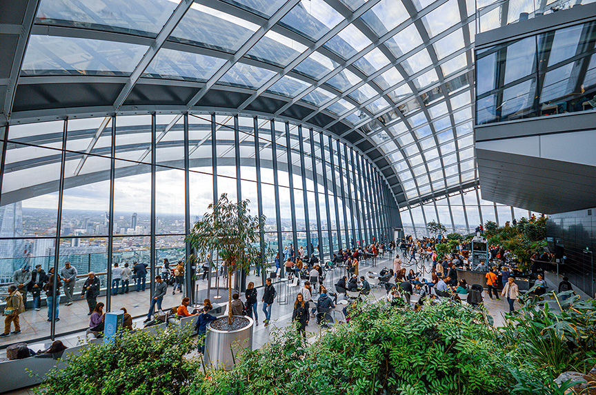 The image depicts a modern, spacious indoor area with large glass windows and a high ceiling, filled with numerous people engaging in various activities. The space features abundant greenery and plants, creating a lively and vibrant atmosphere. It appears to be a public or communal area, possibly a rooftop garden or an observation deck, offering a panoramic view of the city outside.
