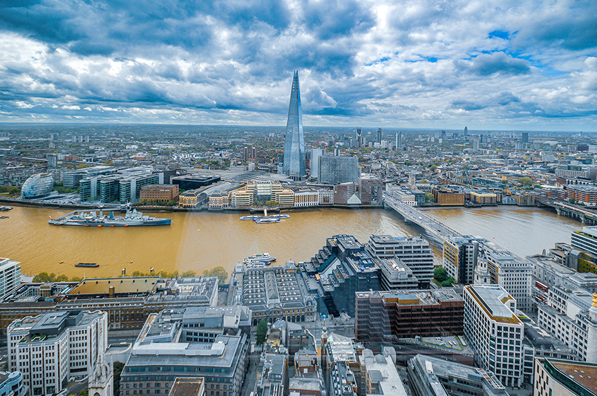 The image showcases a panoramic view of a cityscape with a prominent river running through it. The Shard, a distinctive skyscraper, stands out in the center of the image. The city is densely populated with various buildings, and a large ship is docked on the river. The sky is filled with clouds, suggesting an overcast day. The overall scene captures the bustling urban environment of the city.