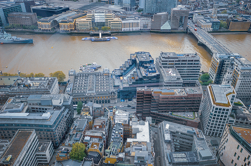 The image depicts an aerial view of a cityscape near a river. The river has several boats and is flanked by buildings on both sides. A bridge spans the river, connecting the two sides of the city. The buildings are a mix of modern and older architecture, with varying heights and designs. The area appears to be a bustling urban environment with a mix of residential, commercial, and possibly industrial structures.