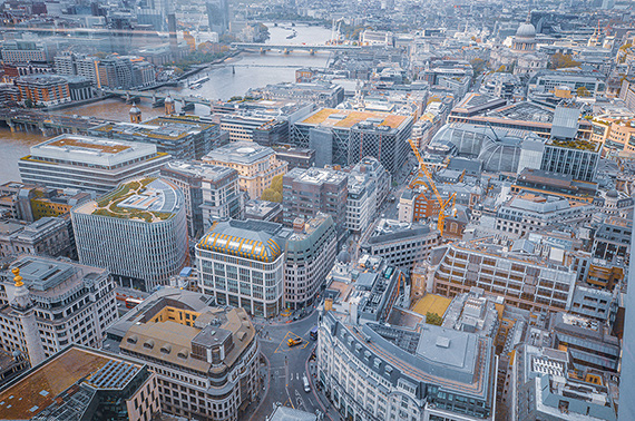 The image depicts an aerial view of a densely built urban area, featuring a mix of modern and older architectural styles. The scene includes a variety of buildings, roads, and a river running through the city. The buildings vary in height and design, with some having distinctive rooftops and others showcasing glass facades. The overall atmosphere suggests a bustling, developed cityscape.