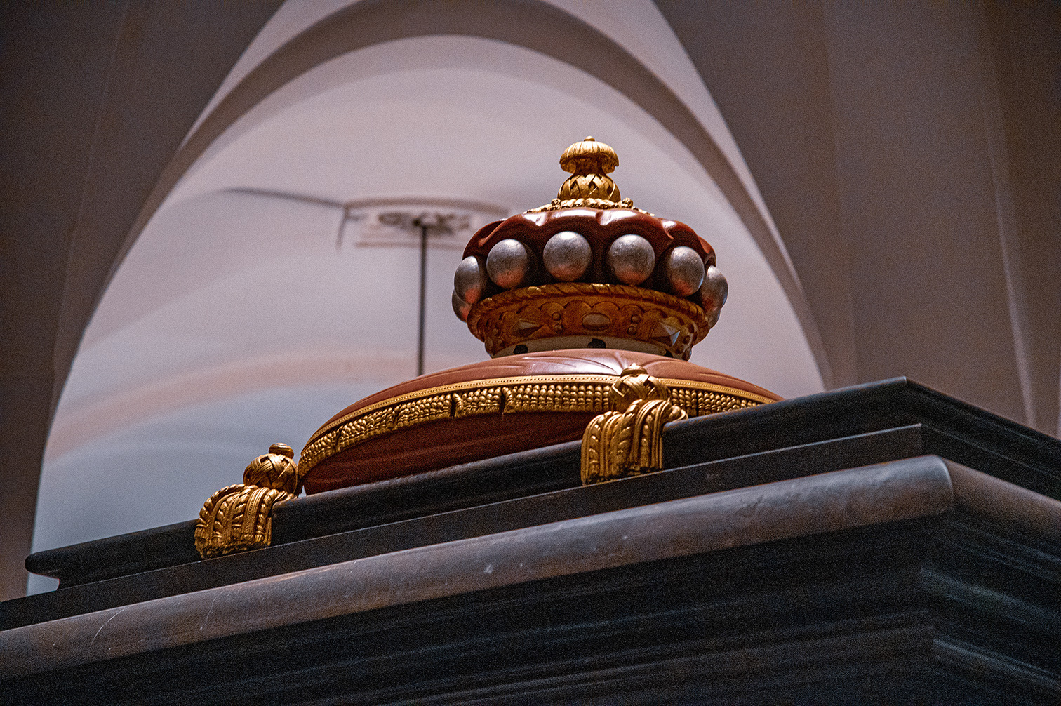 The image depicts an ornate crown, likely a royal or ceremonial one, placed on a pedestal. The crown is richly decorated with gold and possibly precious stones, featuring a prominent arch and intricate detailing. The background includes an arched structure, suggesting the crown is displayed in a museum or exhibition setting.