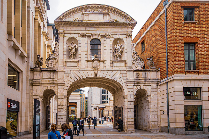 The image depicts an architectural archway in an urban setting, featuring ornate stonework and statues. The archway connects two buildings, with one side showing a store named 'PAUL' and the other side featuring a 'Blacks' store sign. People are walking through the archway, which leads to a pedestrian pathway lined with modern buildings.