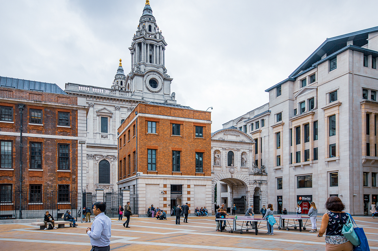 The image depicts a bustling urban square surrounded by historic and modern buildings. The central structure is a prominent church with a tall, ornate tower. People are seen walking, sitting, and engaging in various activities in the open square. The architecture features a mix of brick, stone, and modern materials, reflecting both traditional and contemporary design elements. The overall atmosphere is lively, with a blend of tourists and locals enjoying the space.