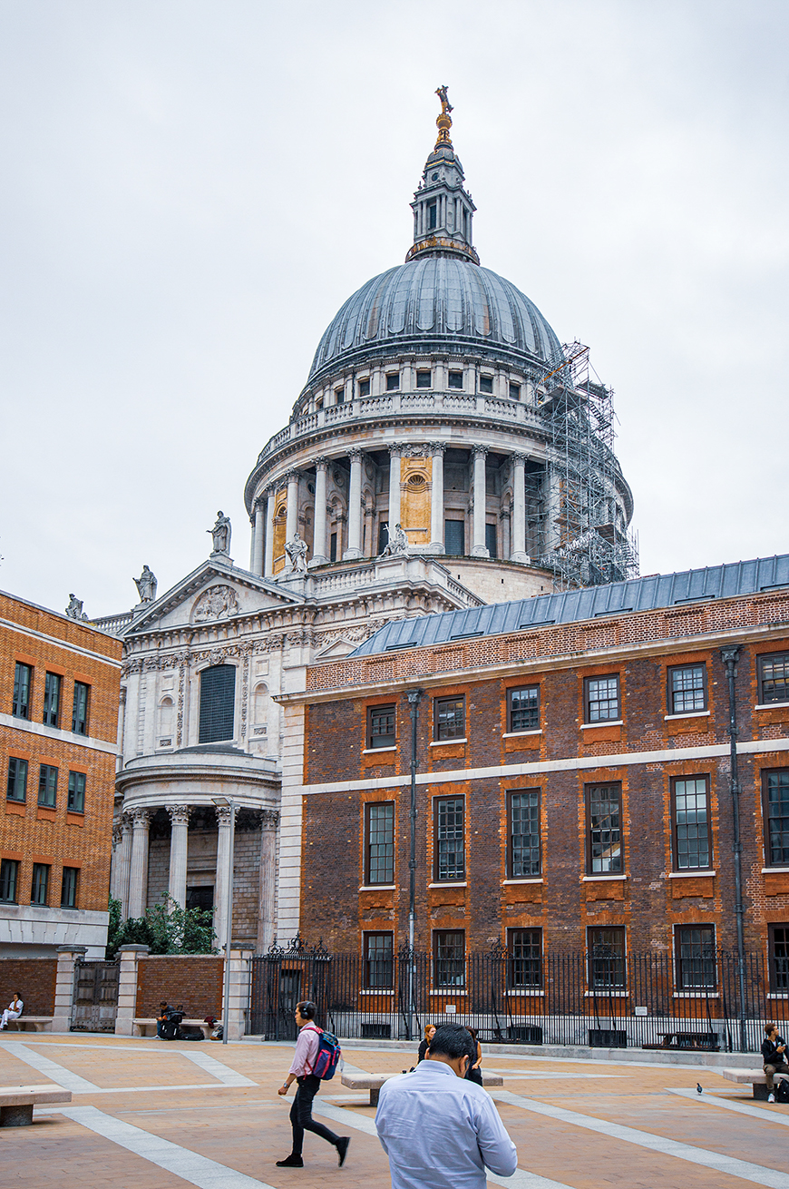 The image depicts St. Paul's Cathedral in London, showcasing its iconic dome and classical architecture. The scene includes people walking and sitting in the foreground, with scaffolding visible on the dome, indicating ongoing maintenance or restoration work.
