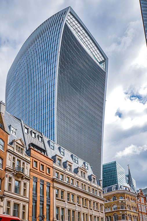 The image showcases a modern skyscraper with a distinctive curved shape, surrounded by older, more traditional buildings. The skyscraper's glass facade reflects the sky, and its unique design stands out against the backdrop of the historic architecture. The scene is set under a partly cloudy sky, highlighting the contrast between contemporary and classical architectural styles.